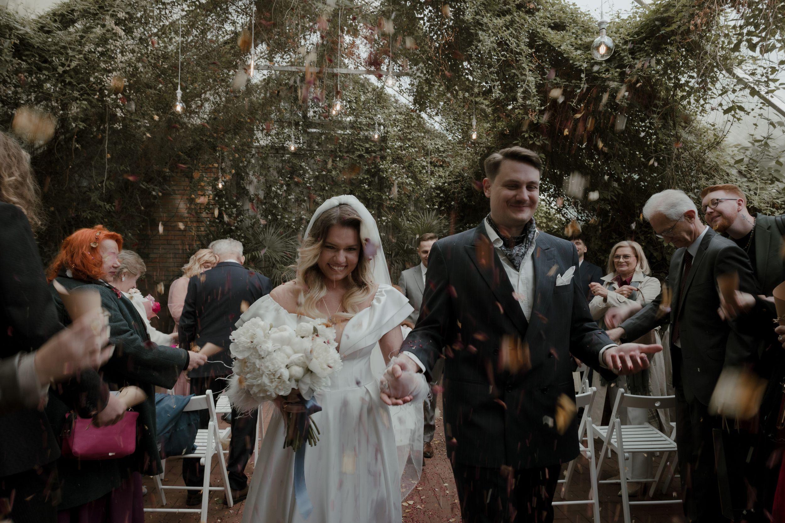 Guests celebrate at an outdoor wedding ceremony under tree canopy with white floral bouquet and formal attire.