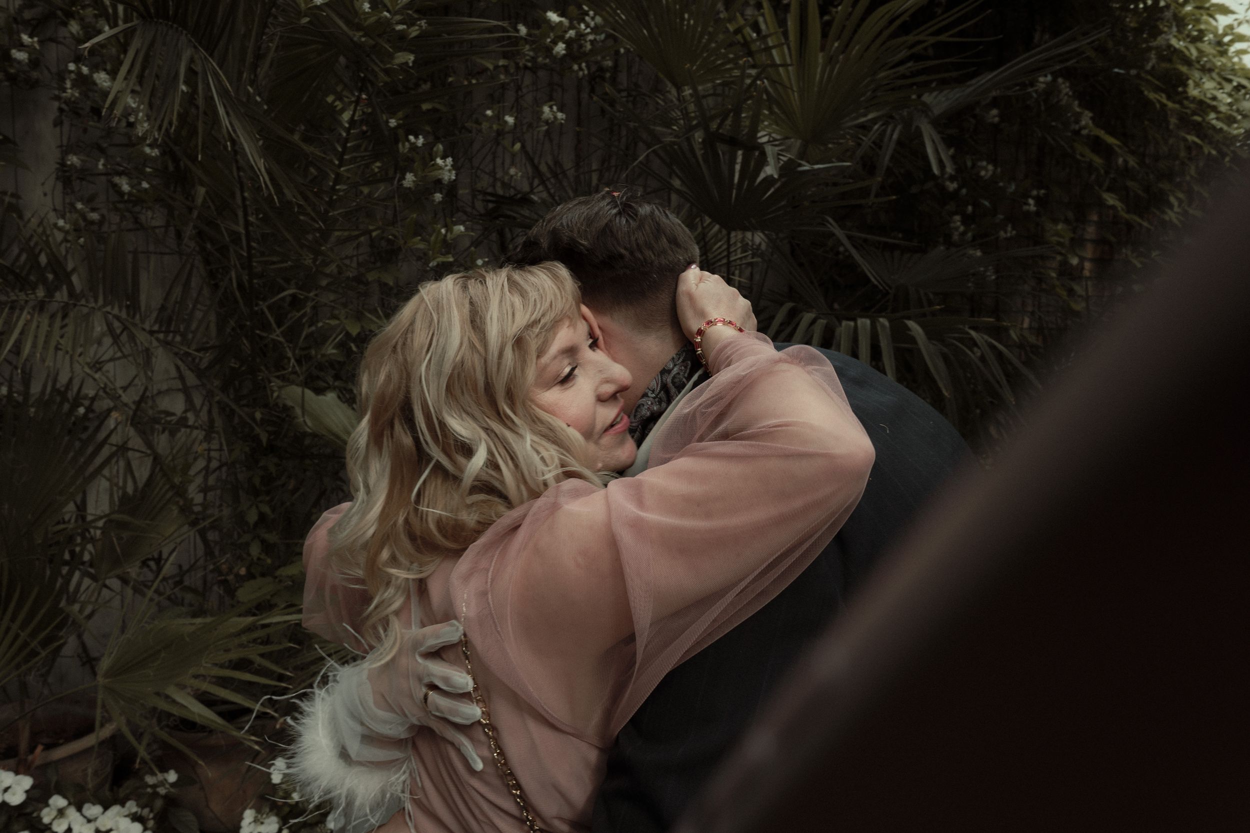 Two people share a tender romantic moment surrounded by lush greenery and trees.