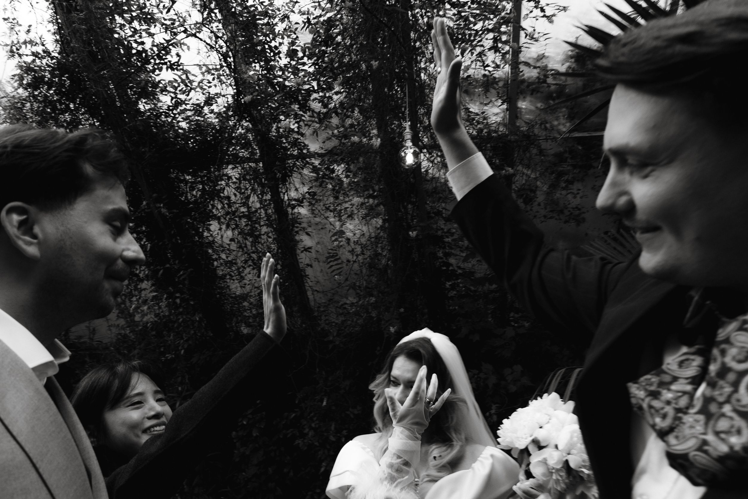 A black and white candid photograph captured during an outdoor wedding ceremony with people gathered in a wooded setting.