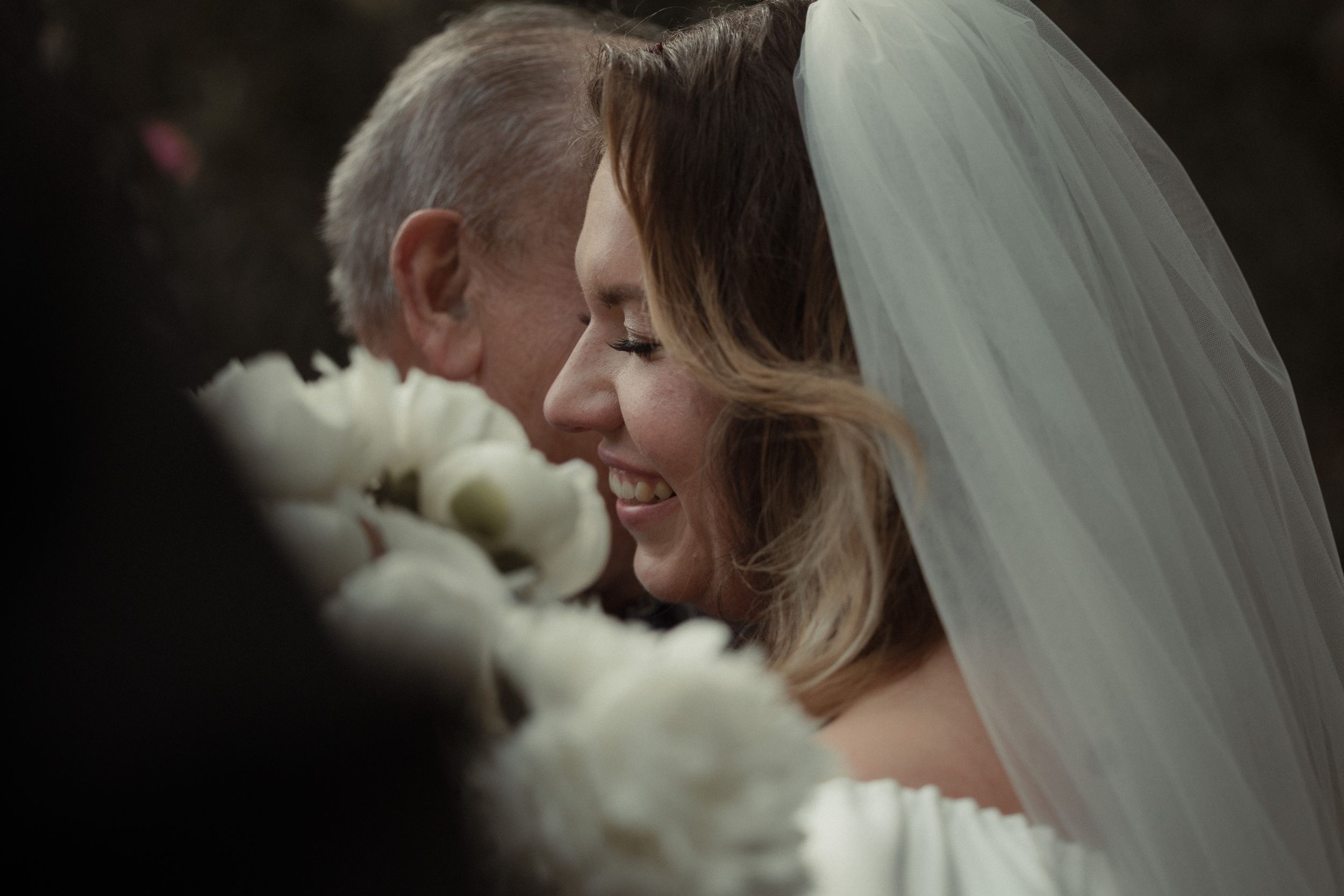 A joyful moment captured at a wedding with white flowers and a flowing veil in a dark, intimate setting.