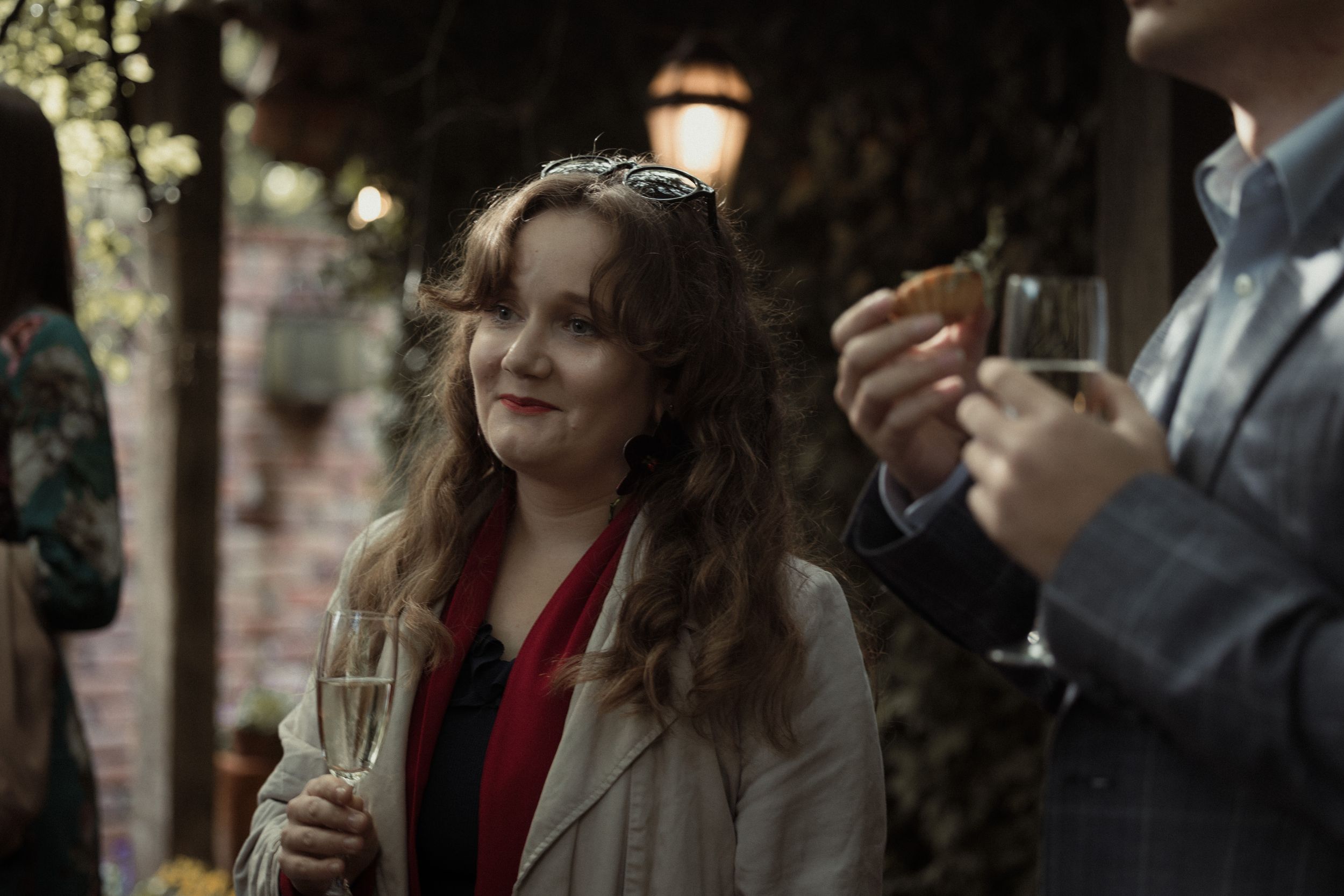 A person with wavy hair in a beige coat and red top holds a champagne glass at an outdoor gathering.