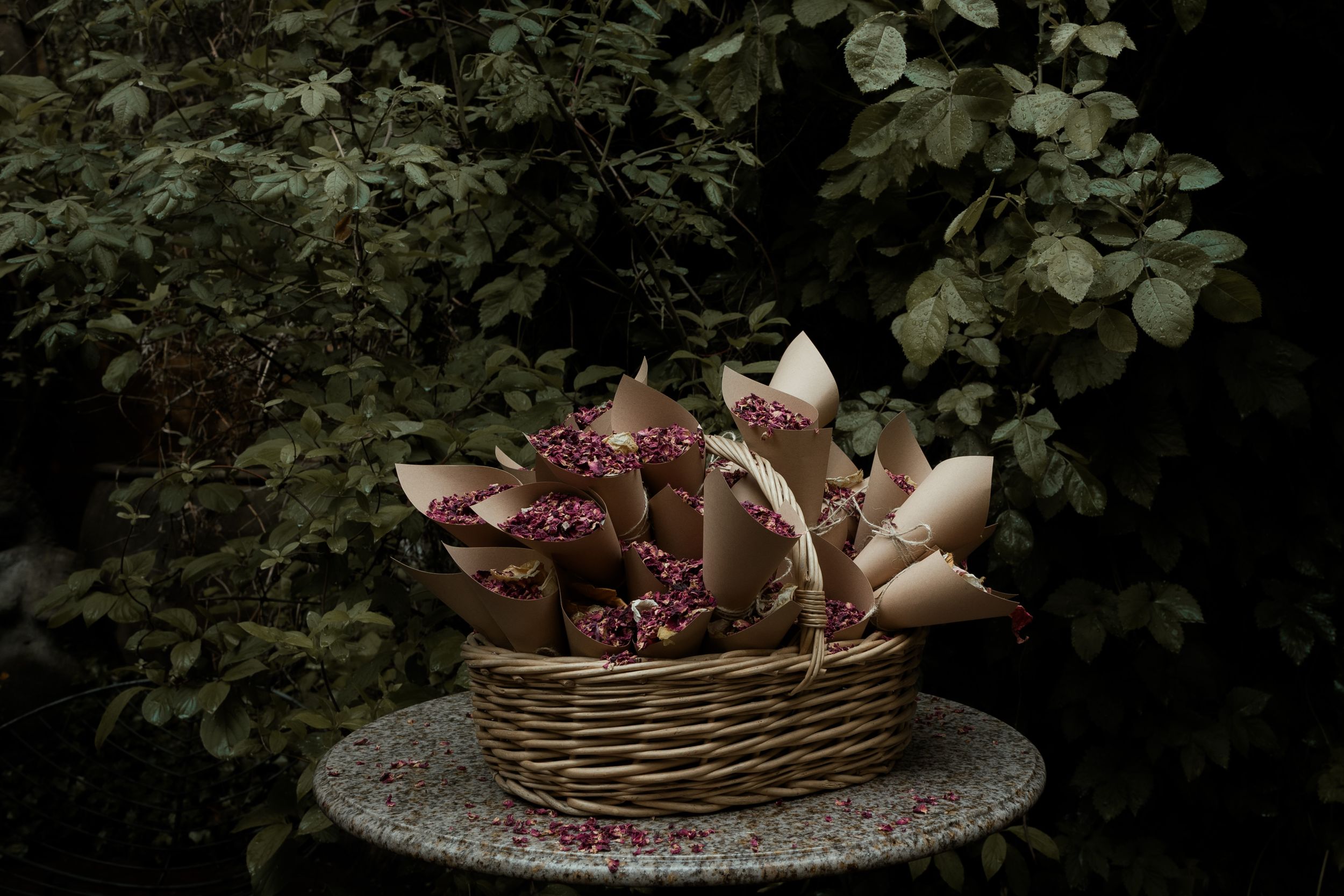 Dark and moody still life arrangement of burgundy and pink flowers in a rustic basket on stone surface against foliage.