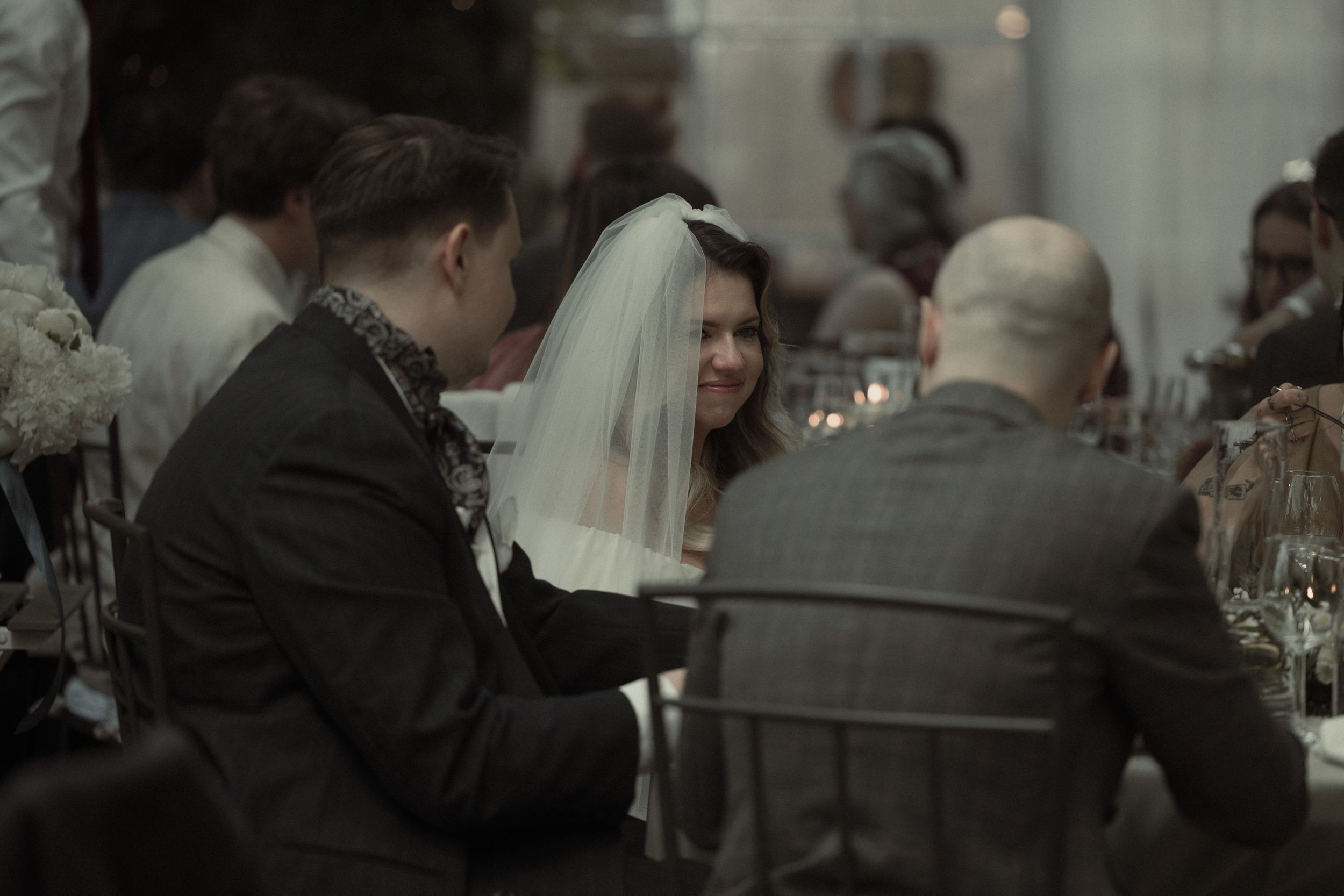 Wedding guests gathered around a dining table in dim lighting with a person wearing a bridal veil among them.