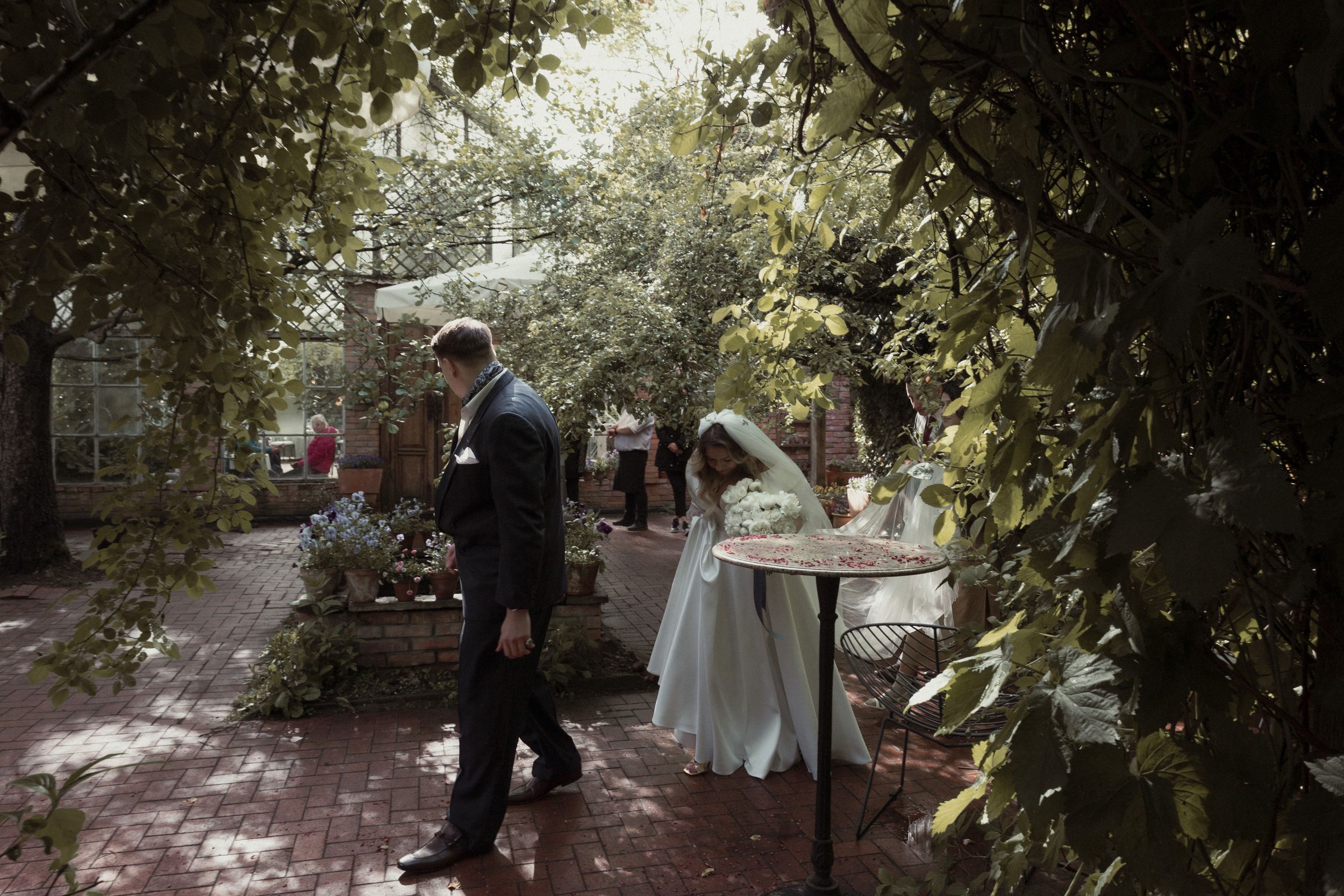 A stone pathway leads through a lush garden with trees and greenery creating a shaded walkway.