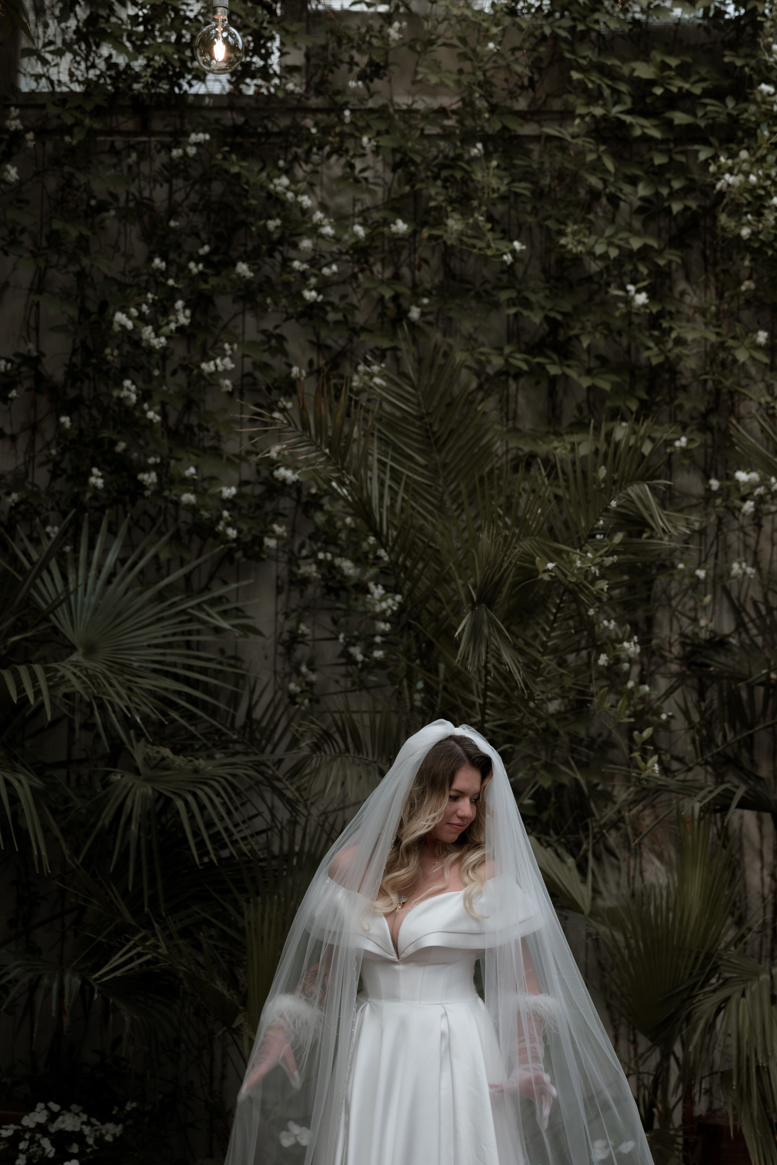 A bride in a white wedding gown and flowing veil stands elegantly against a backdrop of tropical foliage.