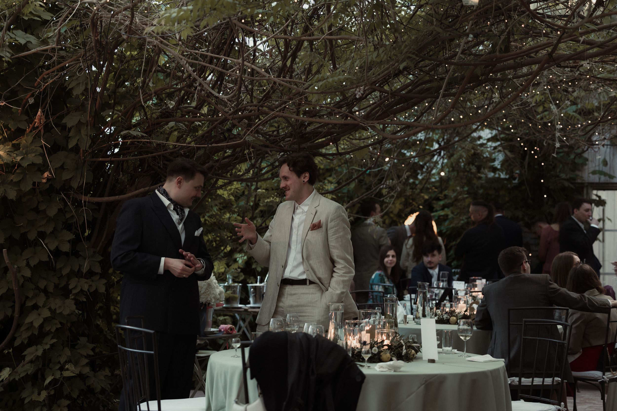 Elegantly dressed guests enjoy a formal outdoor dining event with white tablecloths under a canopy of trees.