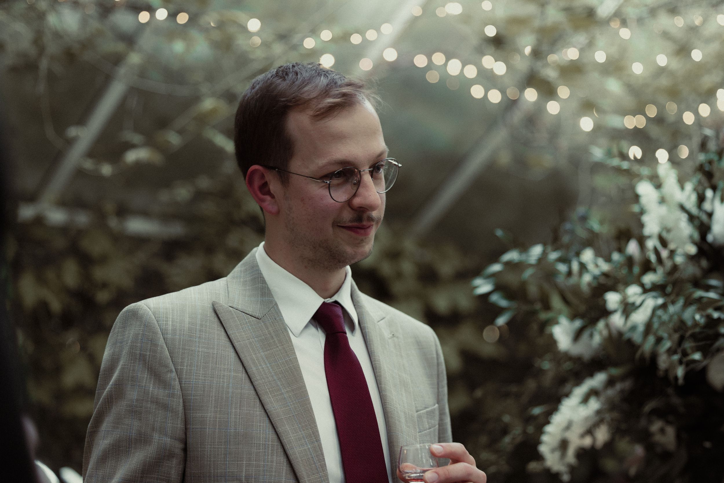 Man in light suit jacket and burgundy tie smiles outdoors with bokeh lighting effect in background.