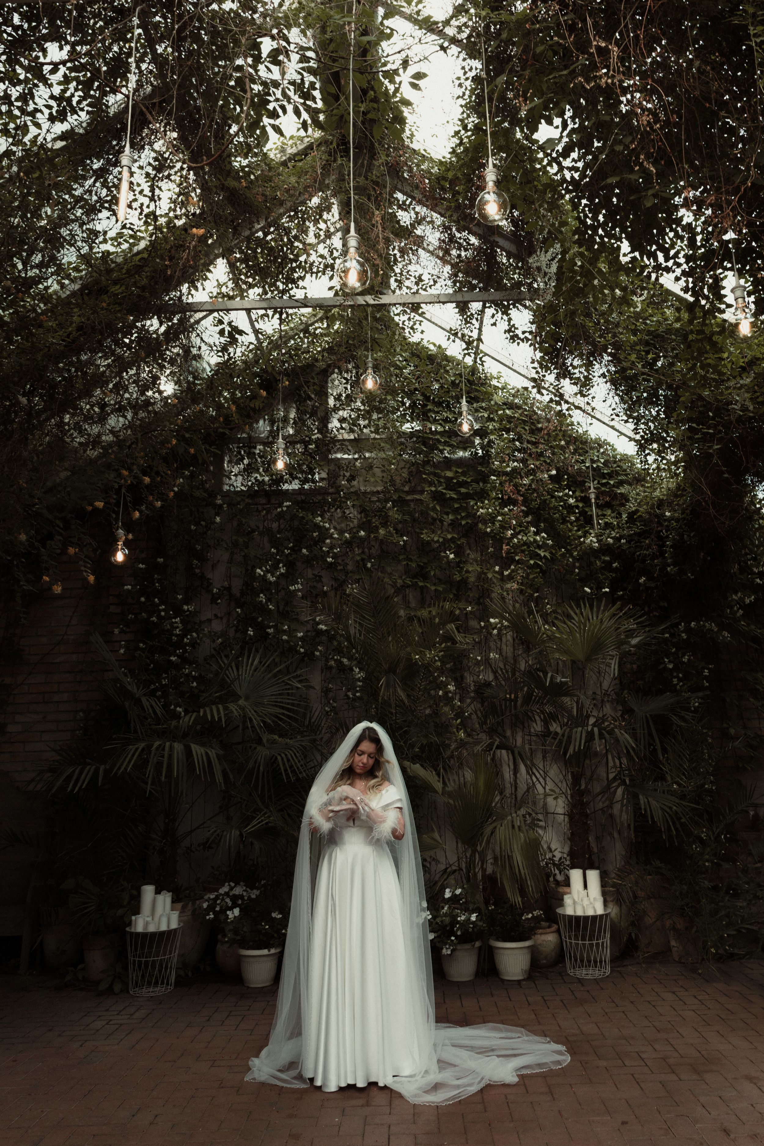 A figure in a white wedding dress with a long veil stands in a dimly lit garden setting under string lights.