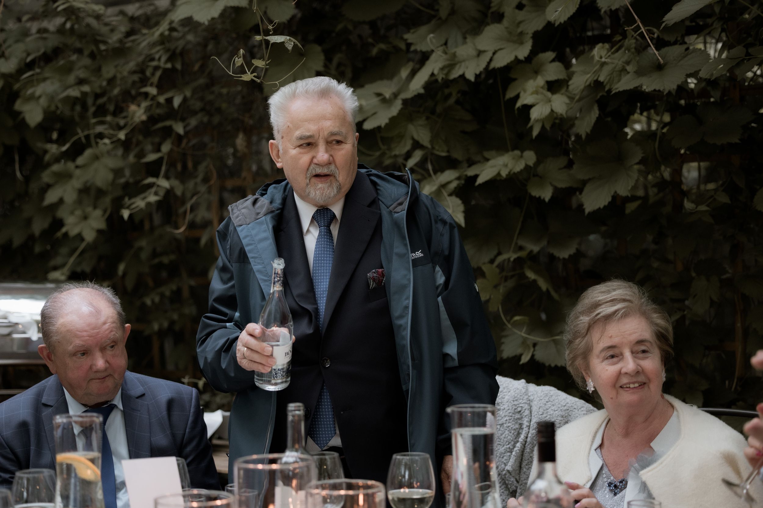 People gathered at an outdoor dining table with glasses and place settings among trees and greenery in the background.