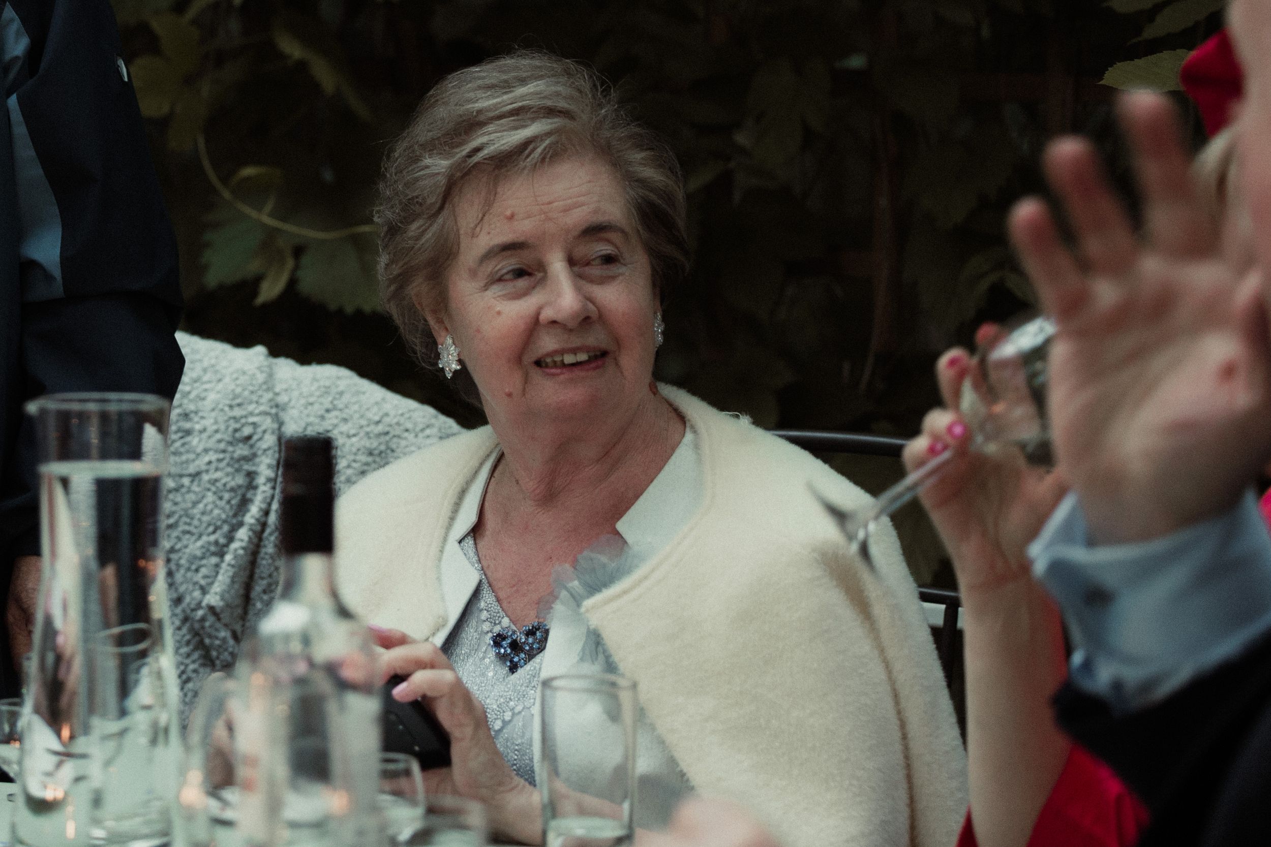 Someone in a cream colored sweater laughs and waves while sitting at a table set for dining outdoors.