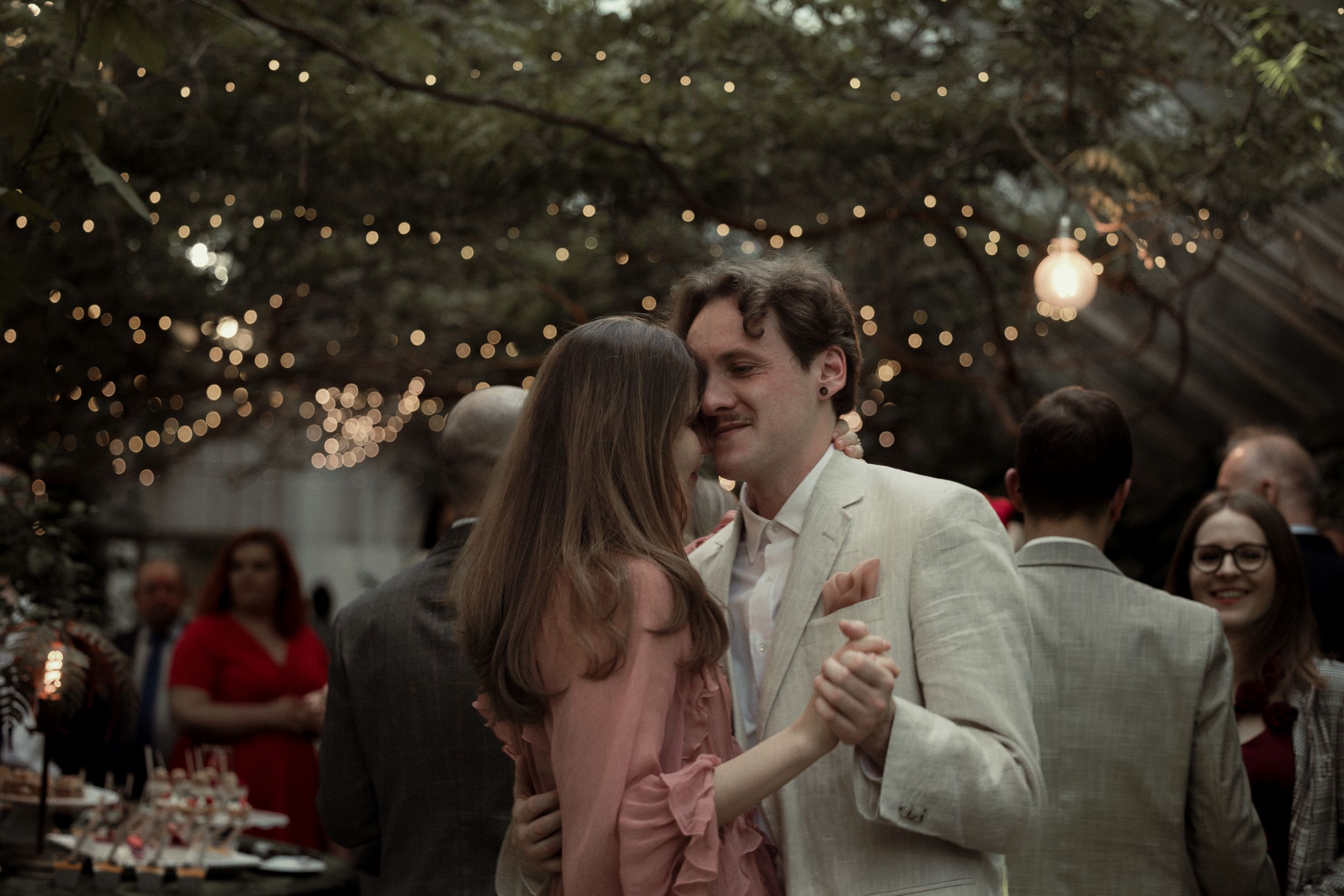 A couple dances together at a romantic outdoor wedding reception with string lights overhead and guests watching.