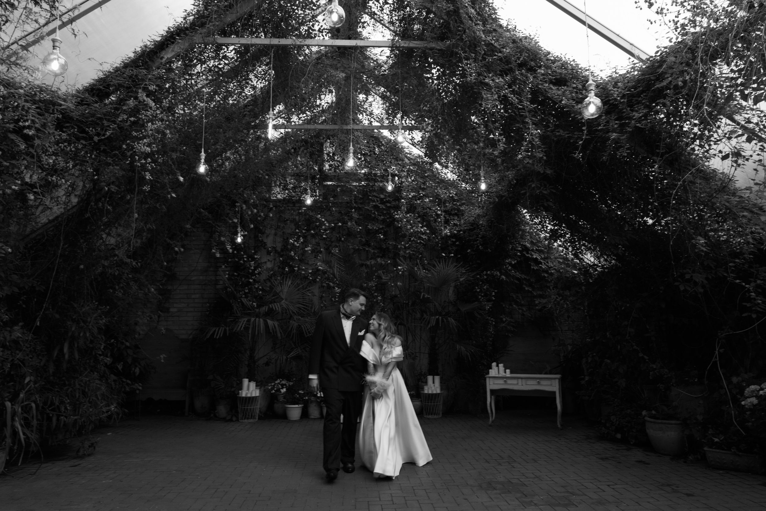 Couple embraces under twinkling string lights in romantic garden venue captured in black and white.