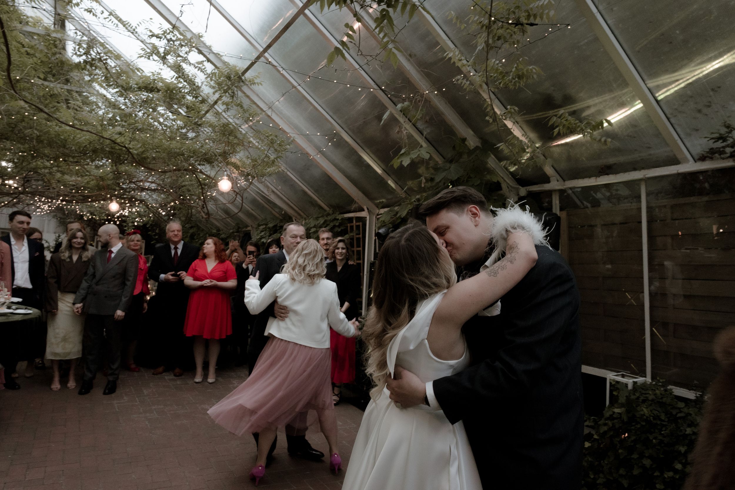 Wedding guests gather around a dance floor inside a rustic greenhouse venue during an evening celebration.