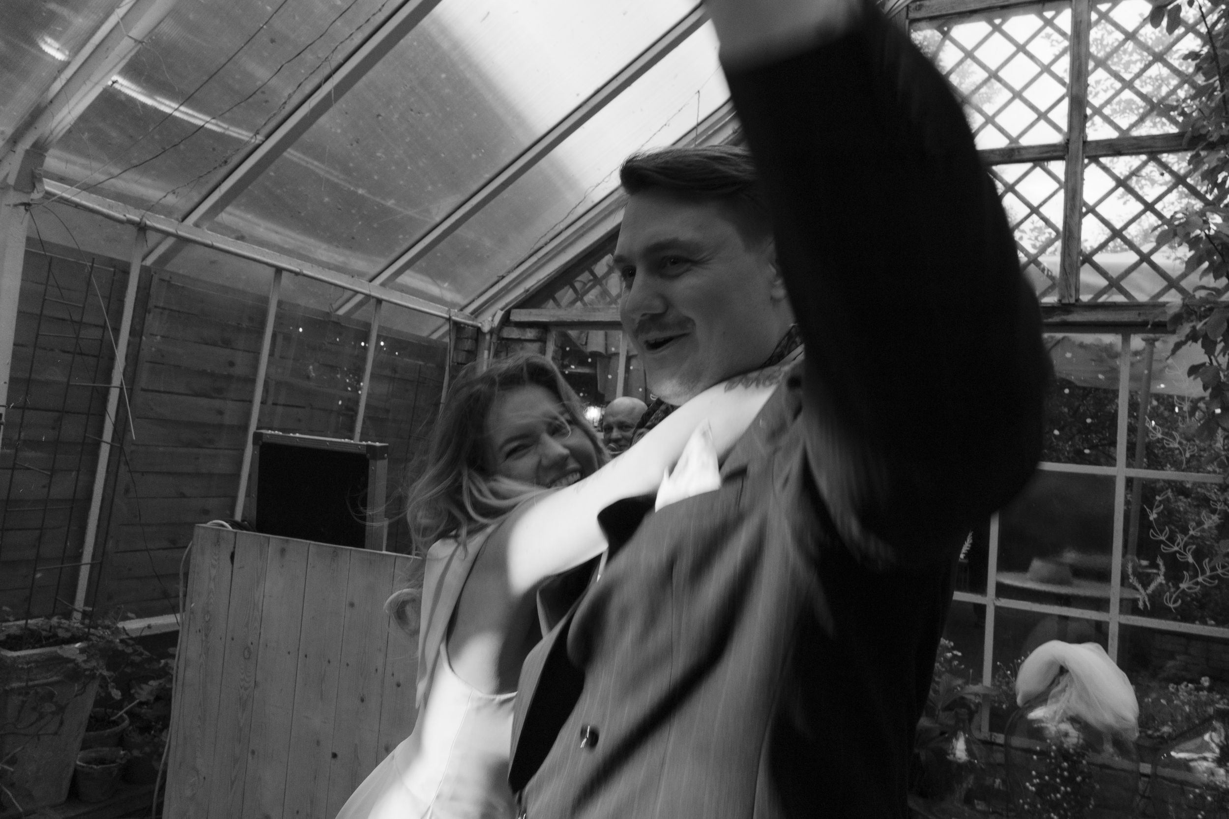 A black and white photograph taken inside a greenhouse shows a figure in a coat making an upward gesture.