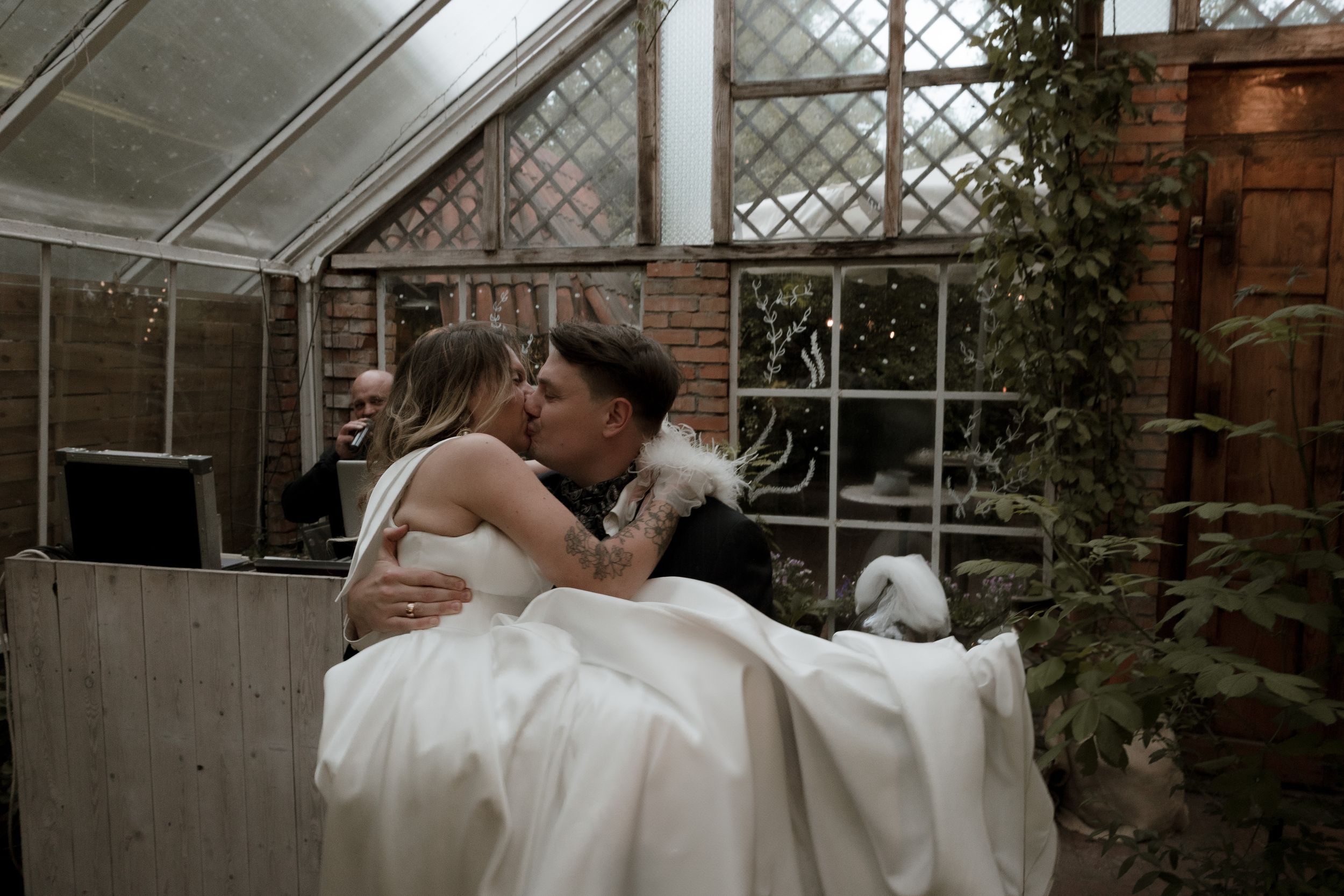 A couple shares a romantic embrace in a greenhouse setting, with the person wearing a flowing white dress.