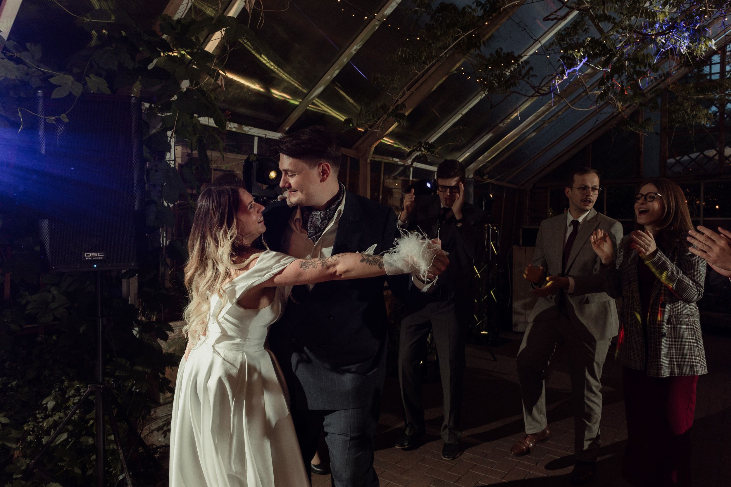Wedding couple dancing under twinkling lights in a rustic indoor venue with guests watching in the background.