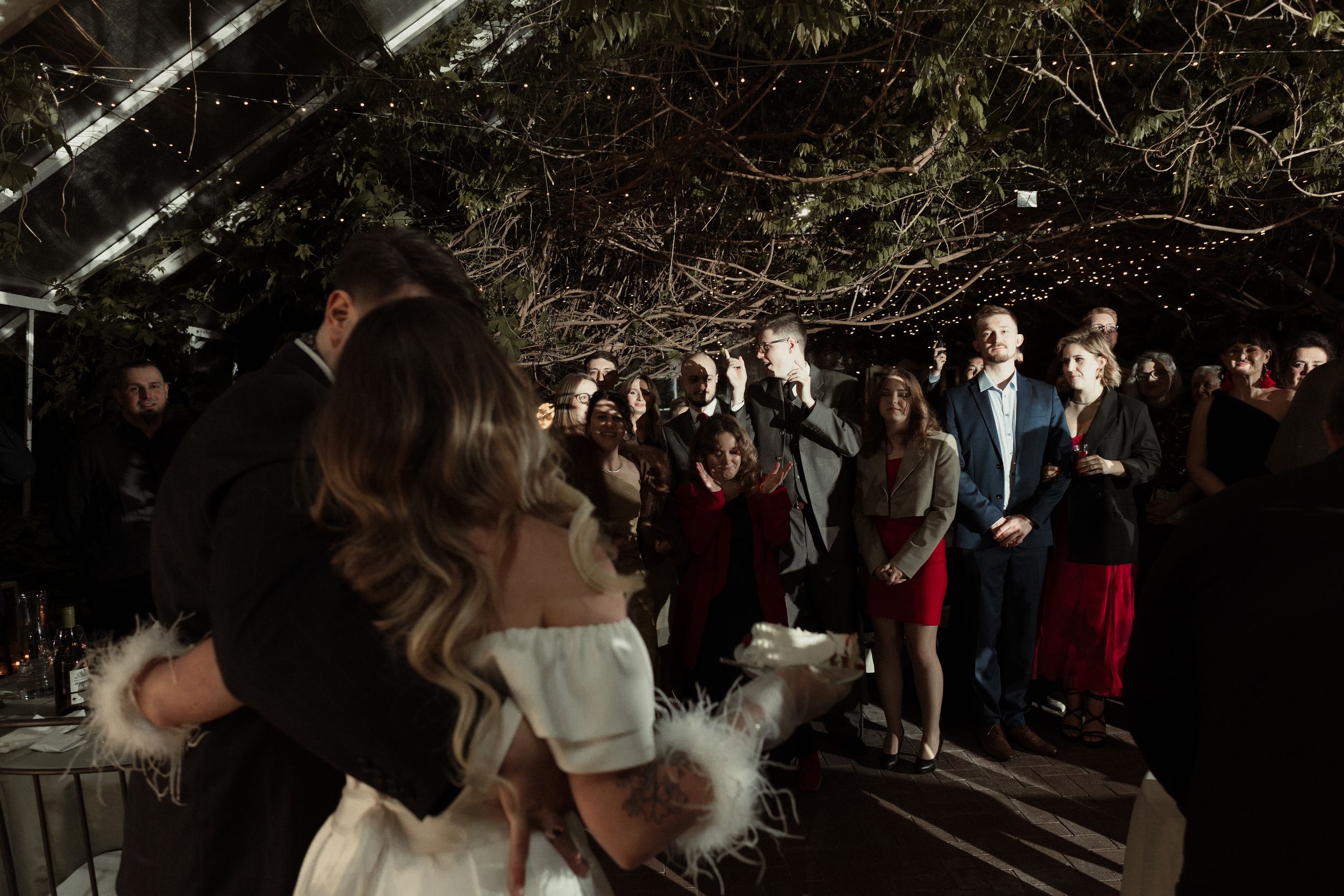 Evening wedding reception with guests dancing under twinkling lights and greenery decorations.