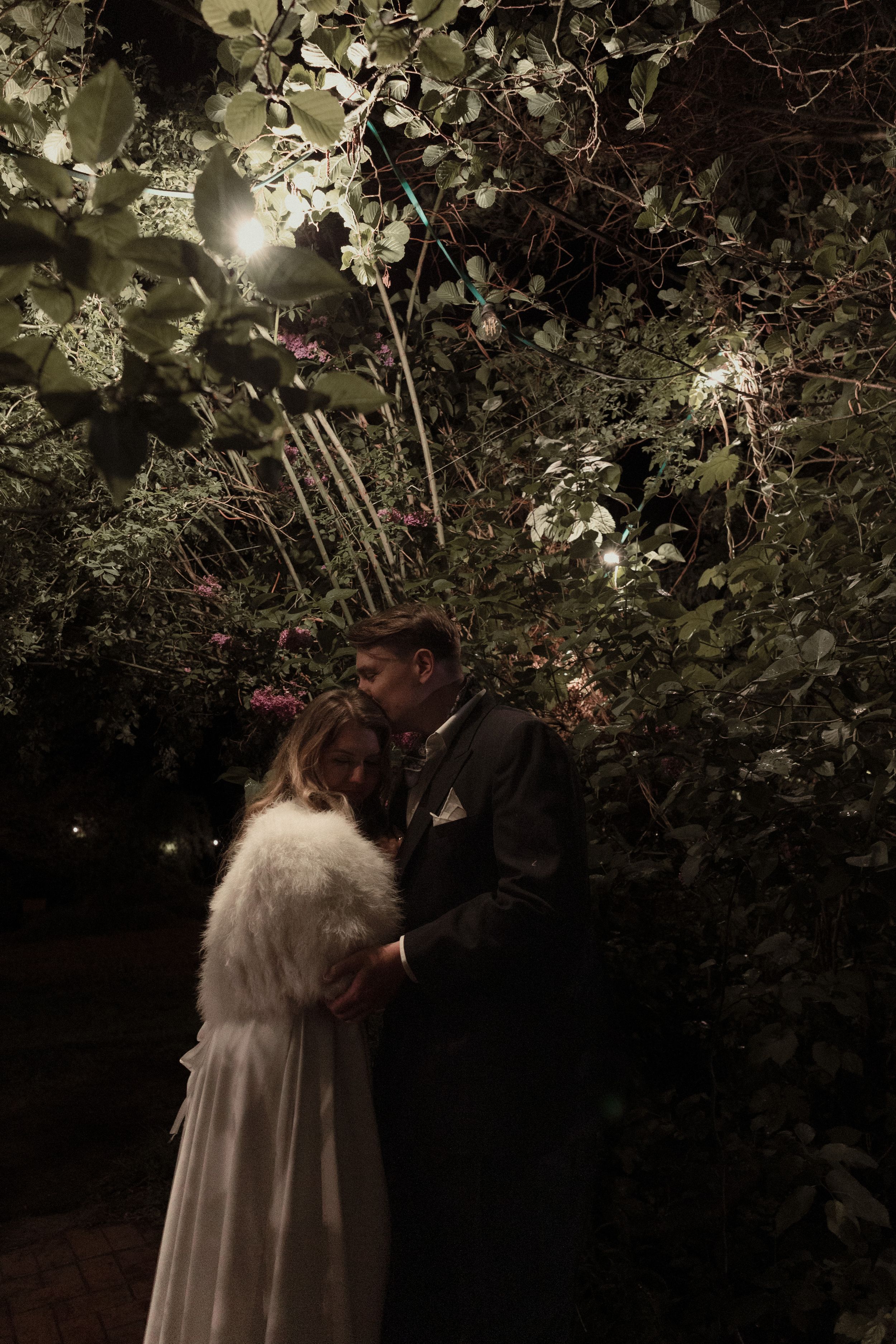 Two people in wedding attire embrace under illuminated cherry blossoms at night.