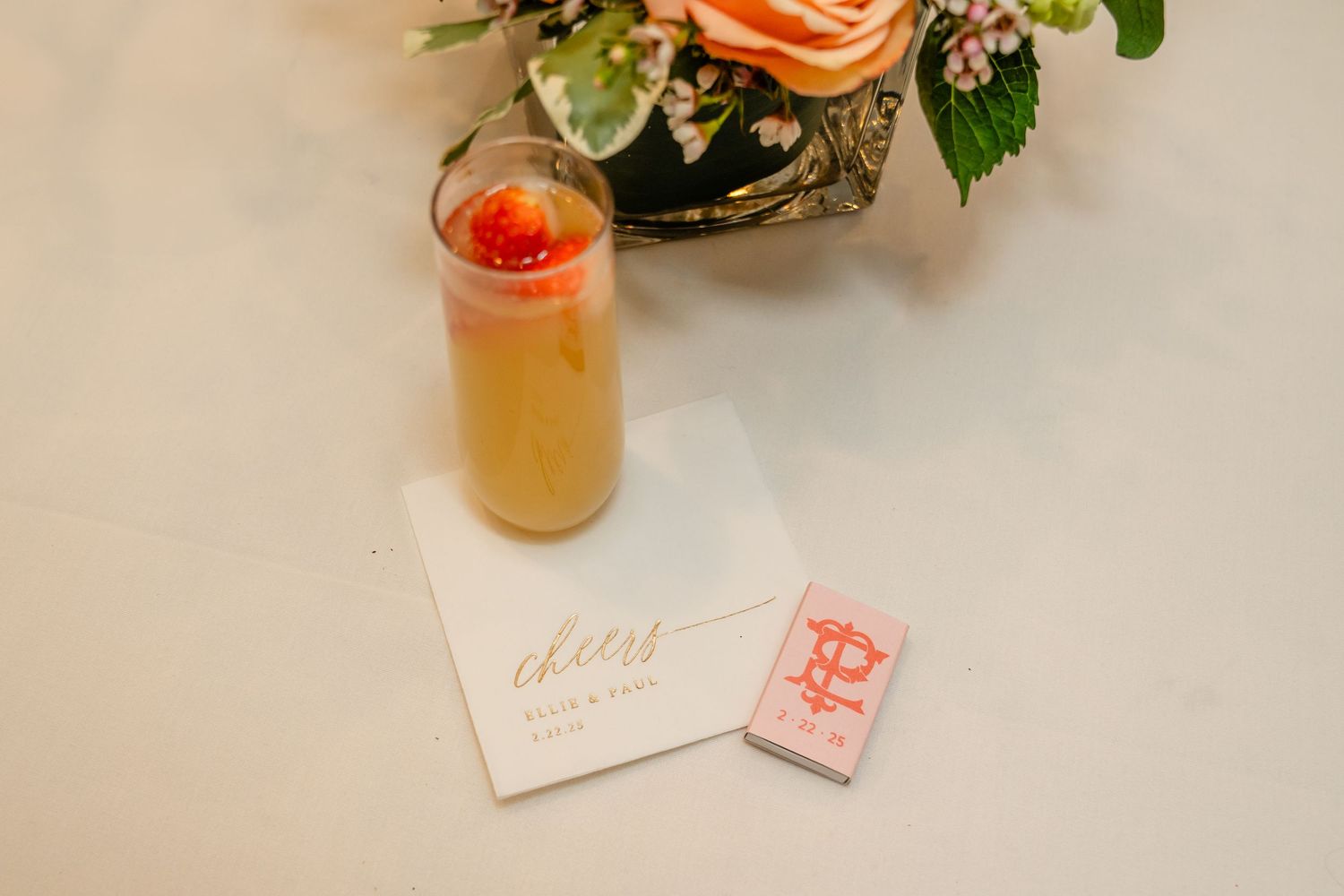 A cocktail with a cherry garnish sits next to floral arrangements and a white paper card on a table.