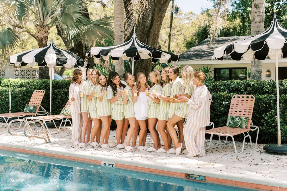 A bridal party in matching light colored pjs stands together near a pool with umbrellas, photo by wedding photographer in Fayetteville NC