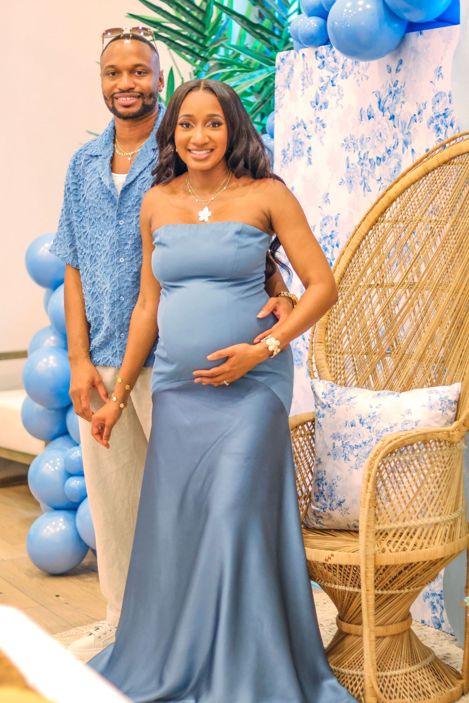 A couple stands together at a baby shower celebration with blue balloons and a wicker peacock chair in the background.