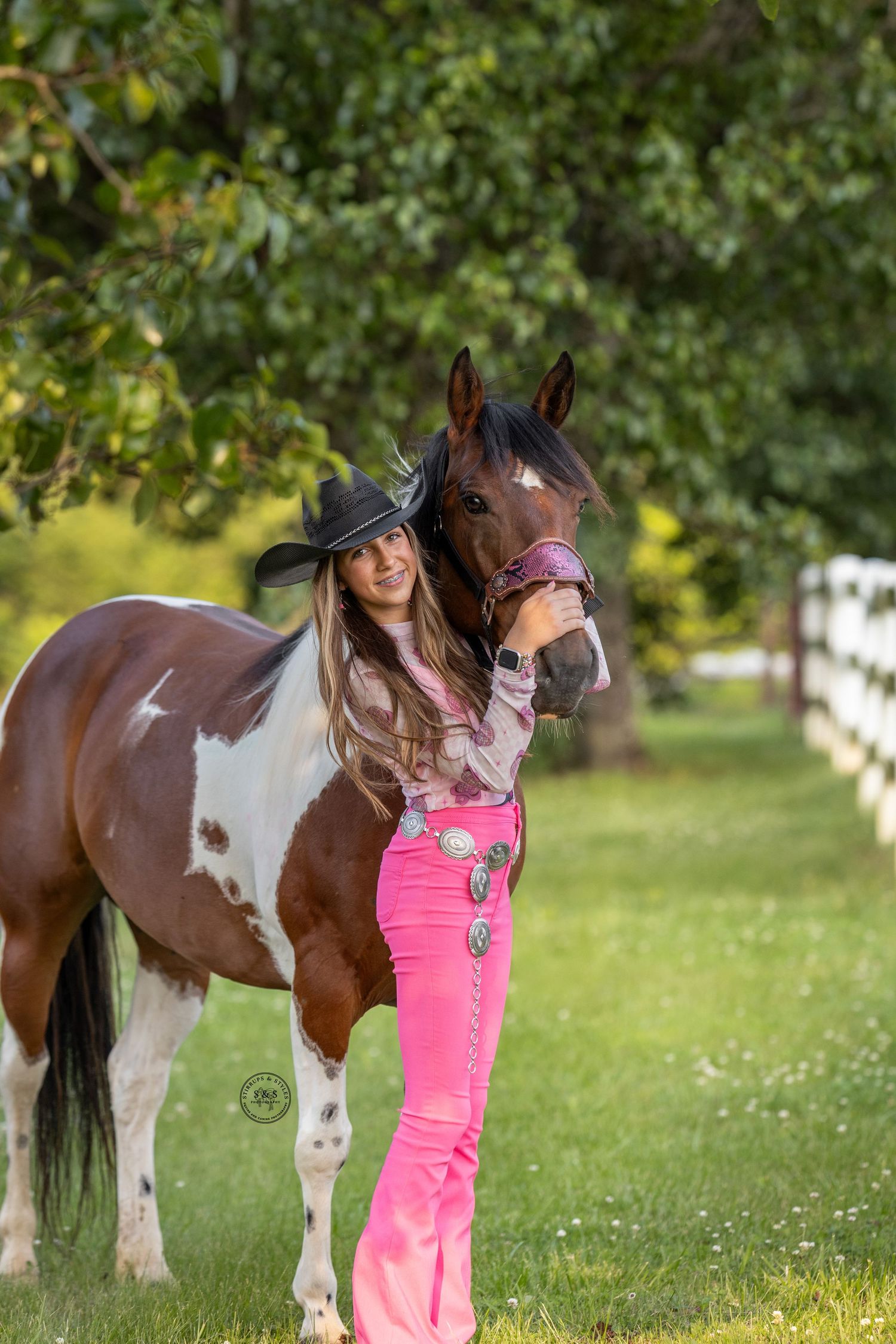 Brown and white paint horse stands in a sunny meadow with someone wearing bright pink beside it.