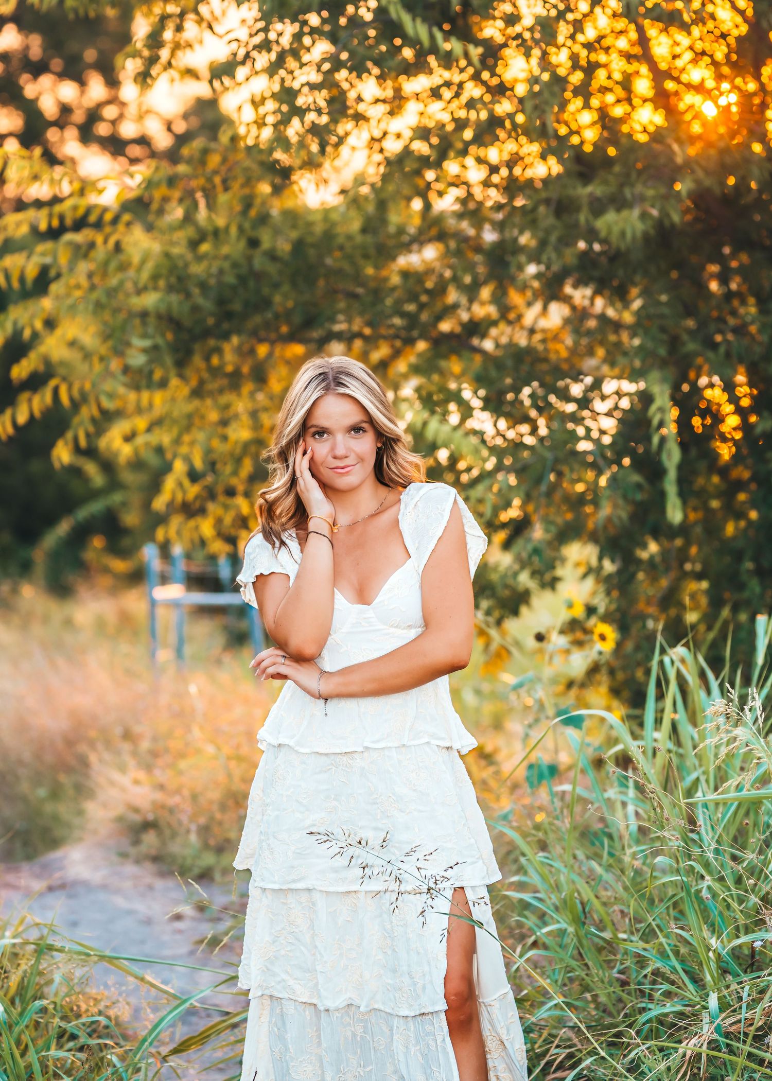 Woman in white dress poses outdoors during golden hour sunset with trees and tall grass in the background.