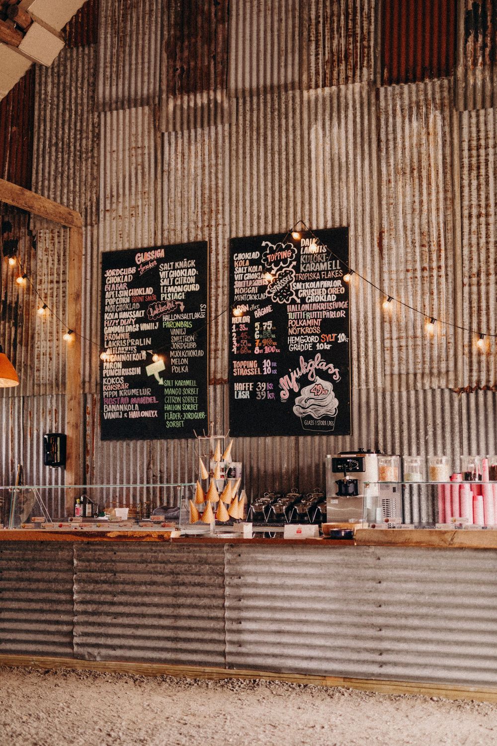 Rustic cafe counter with corrugated metal walls and chalkboard menus displaying drinks and prices under string lights.