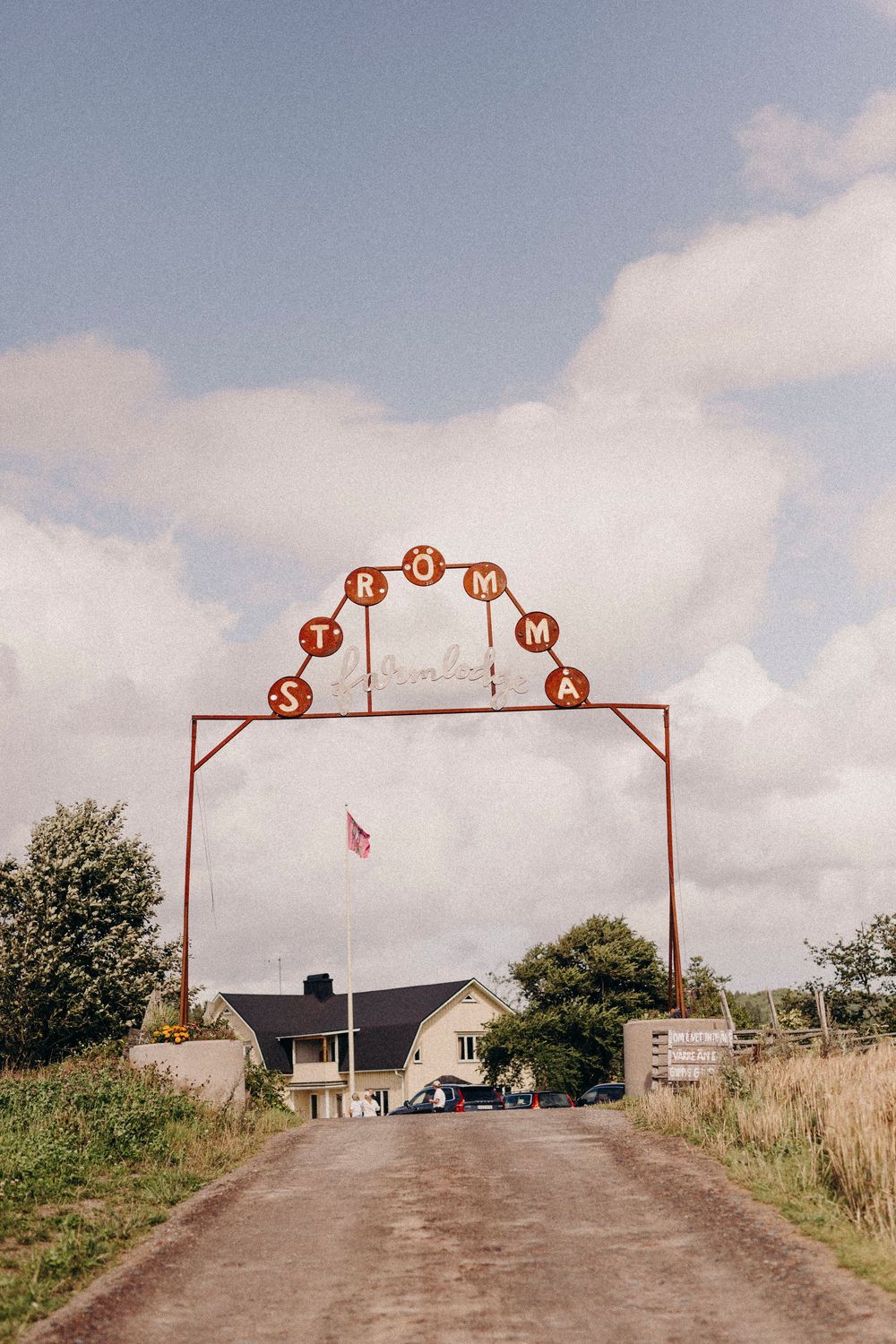A rustic red welcome arch with circular lettering spans a rural dirt road leading to a white farmhouse on a cloudy day.