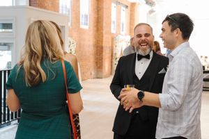 People in formal attire mingle and laugh during a social event in a bright brick-walled venue with natural lighting.
