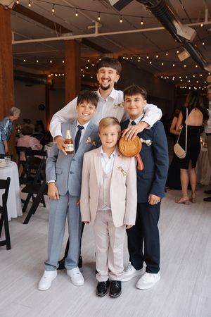 Wedding candid photo of children and groomsmen in suits and formalwear standing on white floor during indoor reception.