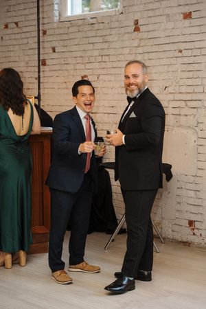Two guests in formal attire share a celebratory moment with drinks at a wedding reception against a white brick wall.