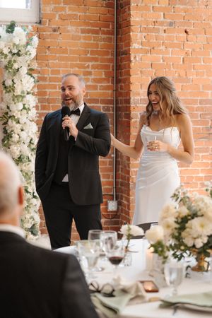Joyful couple shares a laugh during their elegant wedding toast at an industrial brick venue with white floral decor.