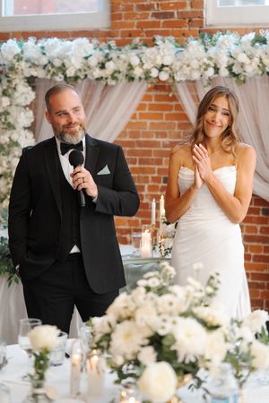 Wedding couple shares an emotional candid moment at reception with white floral decorations against brick wall.