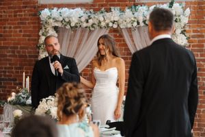 A couple exchanges vows during indoor wedding ceremony with floral arch and brick wall backdrop.