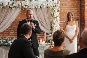 A wedding couple stands in front of a brick wall adorned with white floral and sheer drapery decorations during their ceremony.