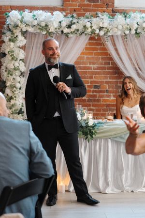 Best man delivers an emotional wedding speech while kneeling in front of guests at a reception venue decorated with flowers.