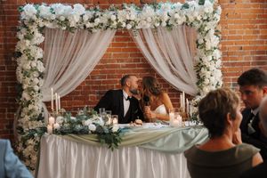 Wedding reception table decorated with white floral arch and sheer draping against exposed brick wall.