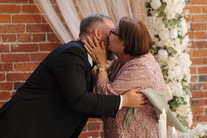 A romantic couple shares a kiss against a brick wall decorated with white floral arrangements.