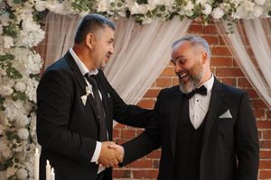 Two people in formal suits greet each other with a handshake against a backdrop of white floral decorations.