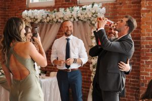 Wedding celebration with drinks in front of floral arch and exposed brick wall at indoor venue.
