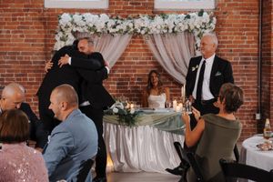 Wedding guests dance and mingle during a reception in a rustic brick-walled venue decorated with floral arrangements.