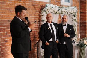 Wedding party in formal black tuxedos and suits gathered near brick wall with white floral decorations.