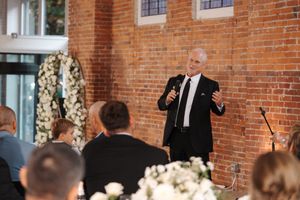 A speaker in a dark suit addresses a wedding reception audience in a rustic brick venue decorated with white flowers.