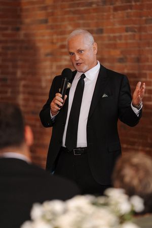 A speaker in a black suit and tie gestures while giving a speech at an event with brick walls in the background.