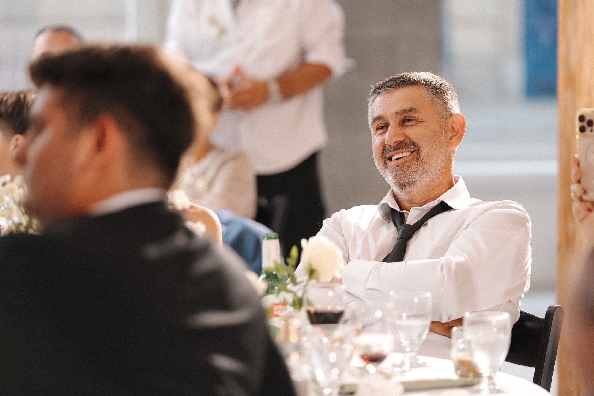 A well-dressed guest smiles during a formal dinner event with wine glasses and table settings in view.