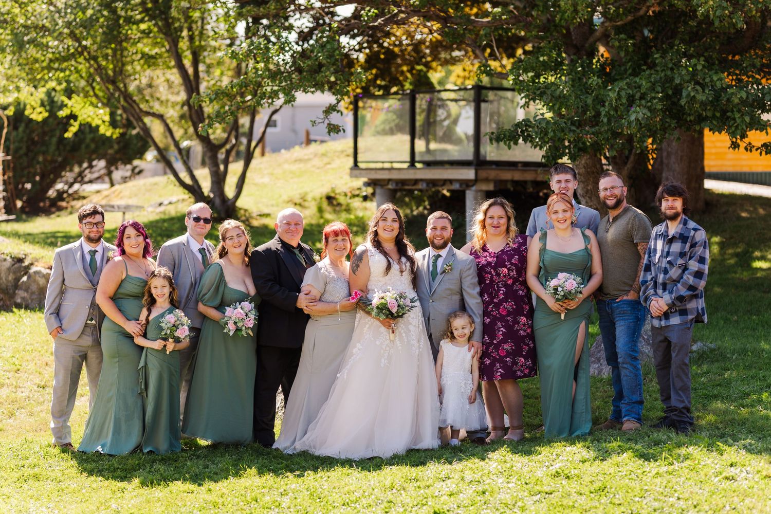 A wedding party standing together on a sunny lawn wearing sage green and gray attire during an outdoor ceremony.