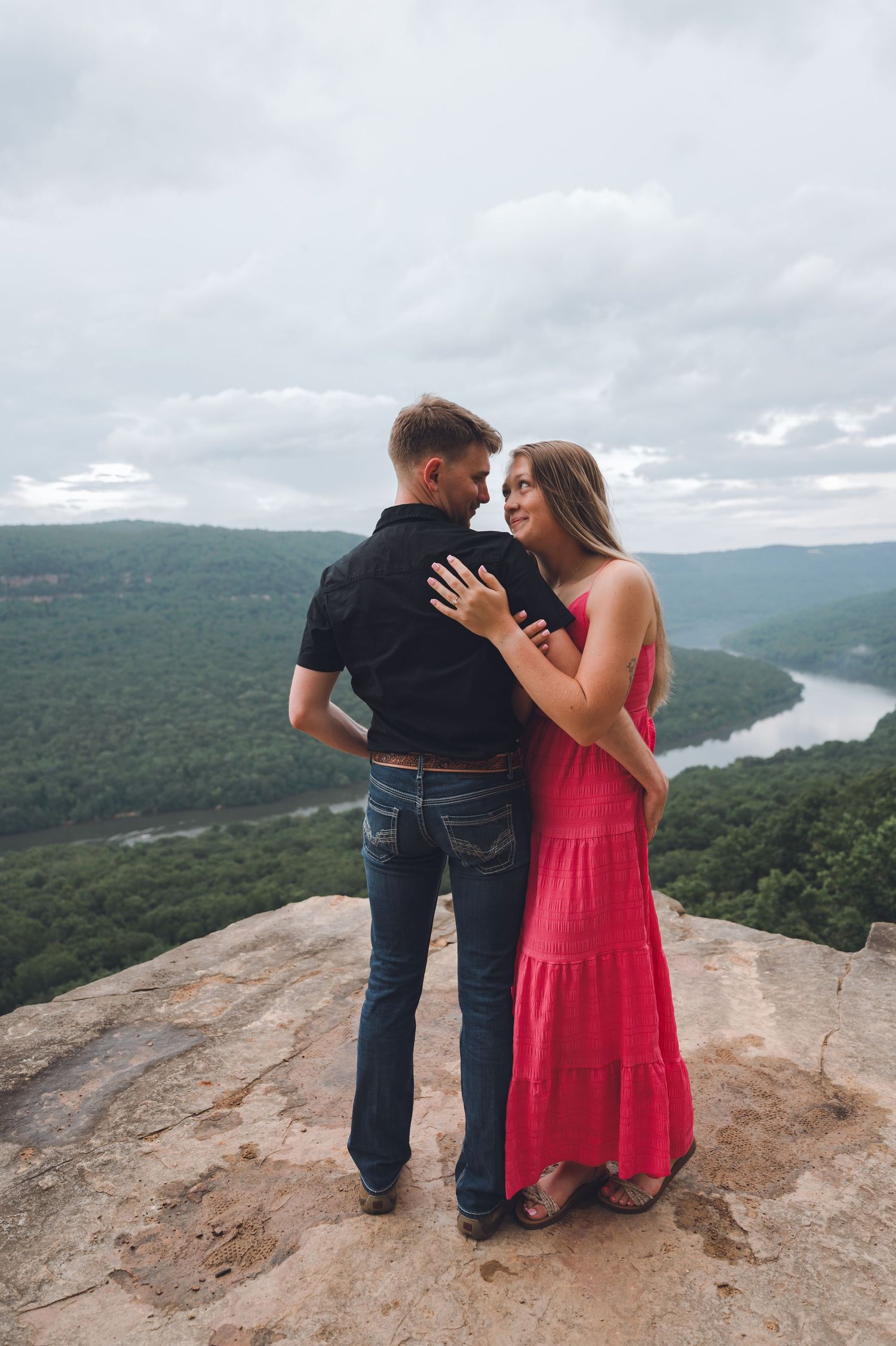 A couple shares a romantic moment atop a scenic mountain overlook with a winding river valley visible in the distance.