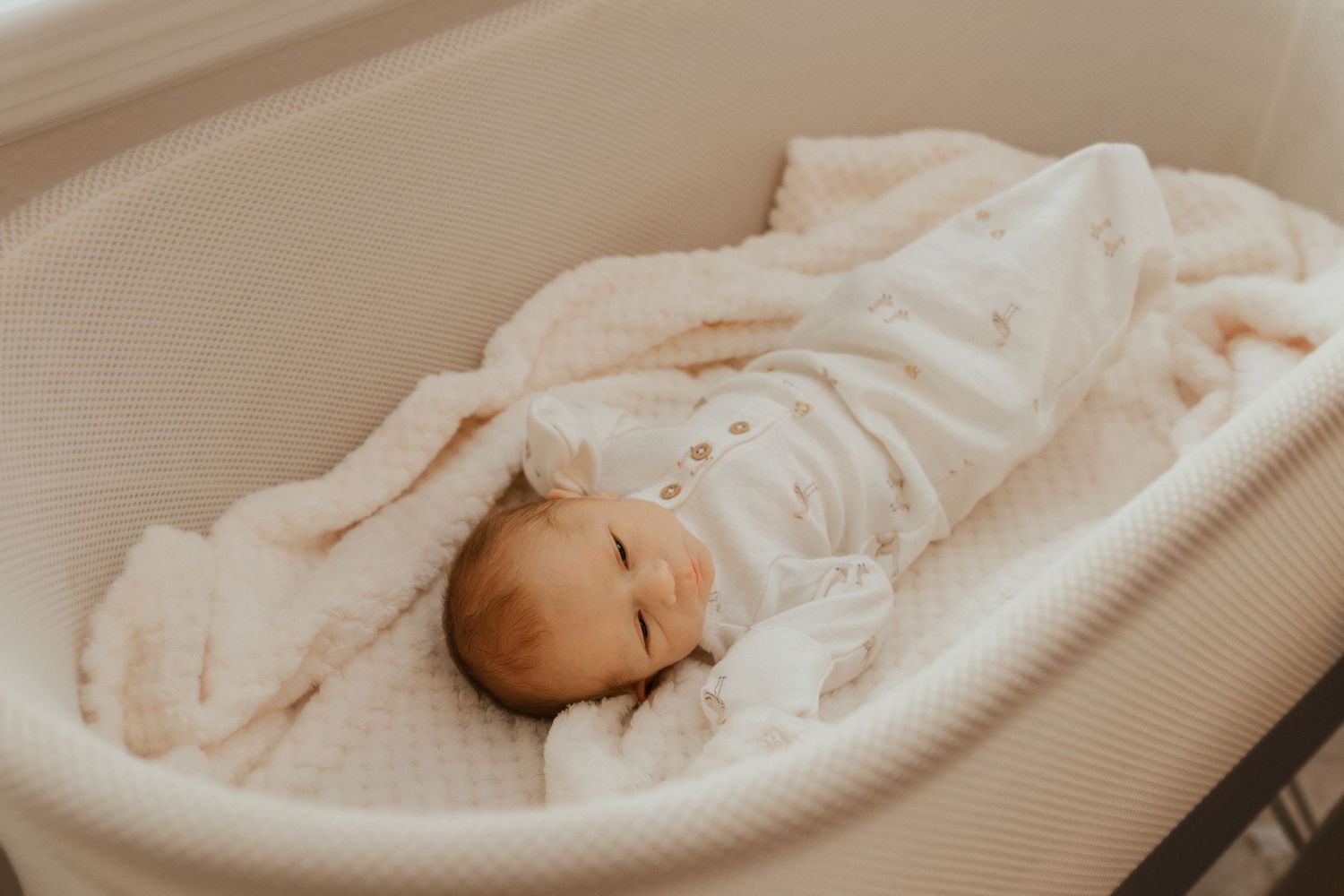 A gentle hand tenderly touches a sleeping infant swaddled in soft white blankets on a beige surface.