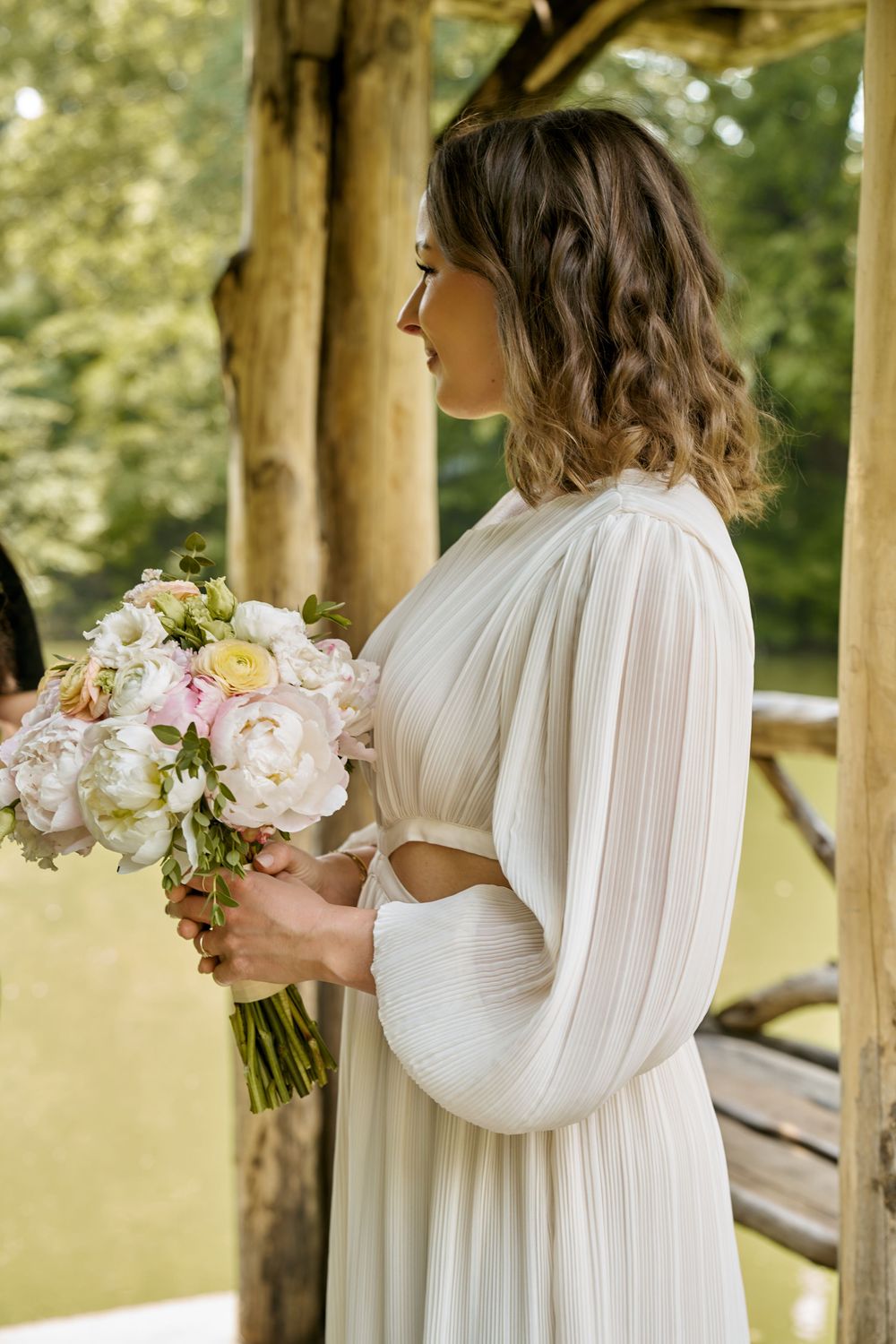 Elegant bride in flowing white dress holds pink and cream floral bouquet in outdoor garden setting. Wedding photoshoot
